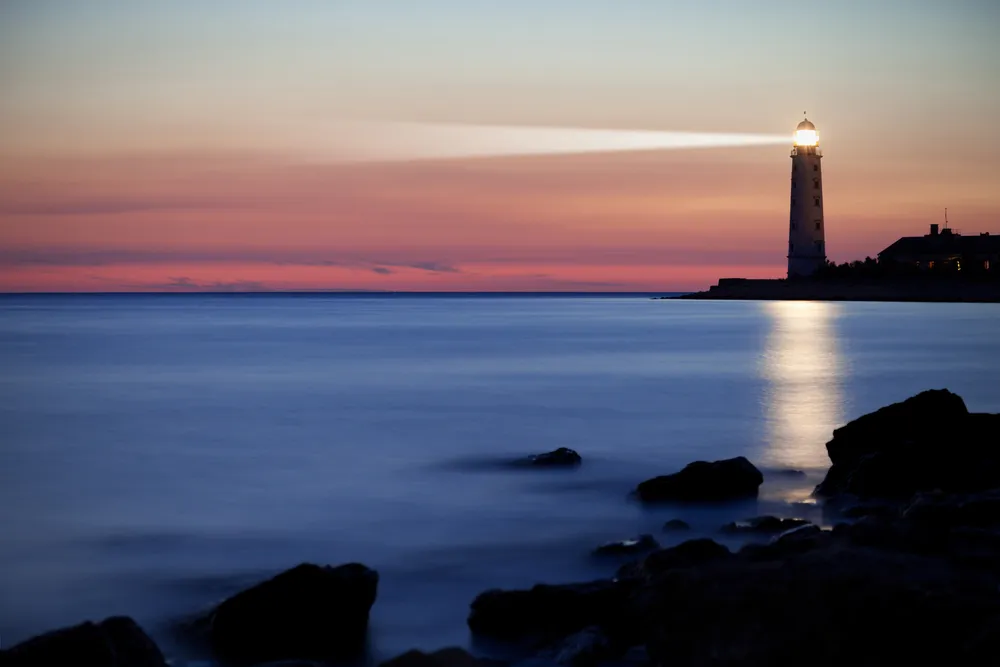 Faro encendido a la orilla del mar al atardecer, con su haz de luz sobre el agua